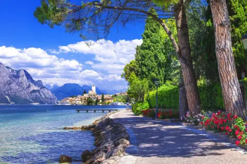 Promenade ensoleillée au bord du lac de Garde, avec des montagnes majestueuses en arrière-plan et un village pittoresque.
