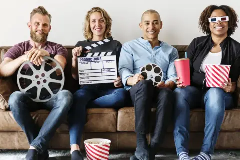 Cztery osoby siedzą na kanapie, trzymając rekwizyty filmowe: klaps, rolki taśmy filmowej, popcorn i napój. Różne rodzaje filmów i ich tworzenie.