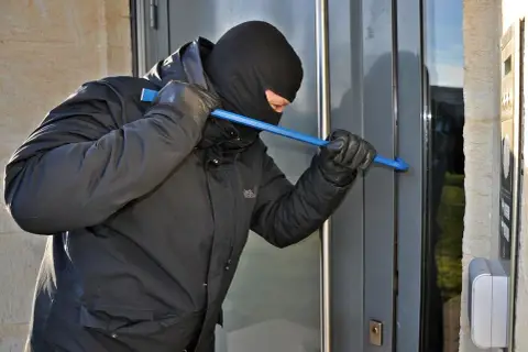Złodziej w kominiarce i rękawiczkach próbuje wyważyć drzwi łomem. To może być próba włamania, zgodnie z art. 279 kk.