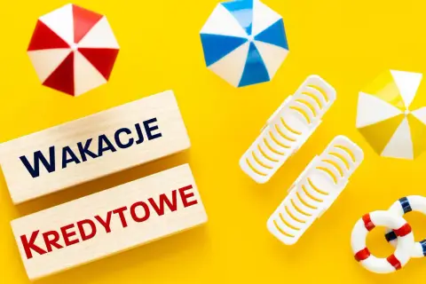 Drewniane klocki z napisem "WAKACJE KREDYTOWE" na żółtym tle, otoczone miniaturowymi parasolkami plażowymi i kołami ratunkowymi.