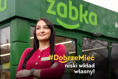 Uśmiechnięta franczyzobiorczyni Żabki przed sklepem. Hasło: #DobrzeMieć niski wkład własny!