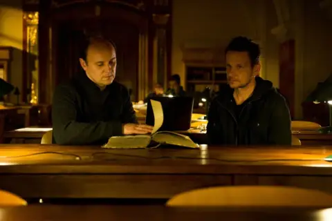 Dwóch mężczyzn przy stole w bibliotece, jeden czyta starą księgę. Scena z filmu "Sługi Boże".