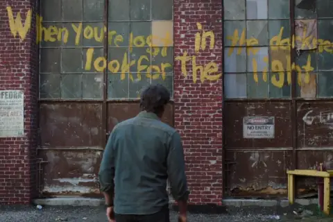 Joel patrzy na napis "When you're lost in the darkness, look for the light" na ścianie budynku, nawiązanie do świetlików z The Last of Us.