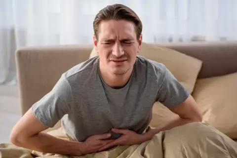 Mężczyzna w łóżku trzymający się za brzuch z wyrazem bólu na twarzy.