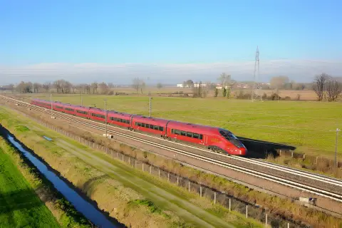 Le train rouge Italo file sur les voies, reliant Roma à Naples à travers la campagne verdoyante sous un ciel bleu.