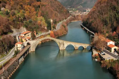 Il Ponte della Maddalena, con i suoi archi irregolari, attraversa un fiume circondato da colline autunnali e case colorate.