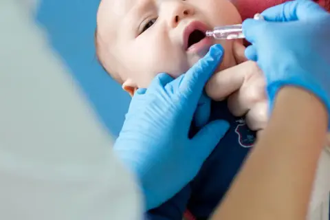 Ein Baby erhält Tropfen zur Vorbeugung von Rotavirus-Symptomen. Blaue Handschuhe schützen die Hände des Arztes.