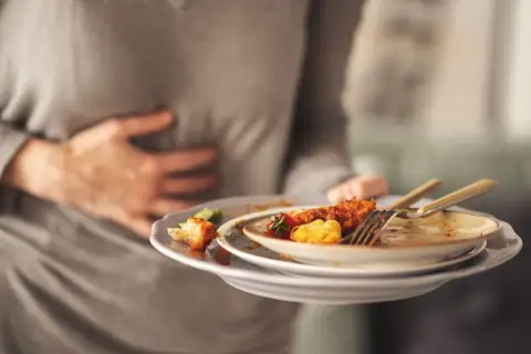 Osoba trzymająca brudne talerze po jedzeniu, masująca bolący brzuch. Dlaczego po zjedzeniu wieprzowiny boli brzuch? Najczęstsze przyczyny.