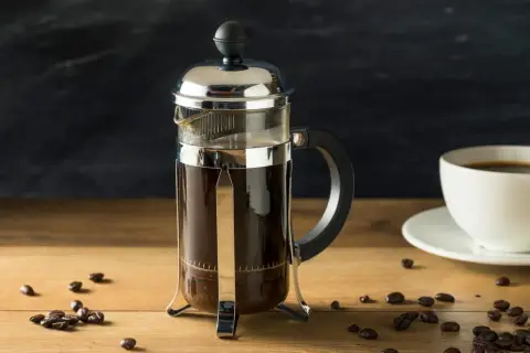Szklany french press z ciemną kawą, obok biała filiżanka i ziarna kawy. Idealny poranek z kawą do french press.