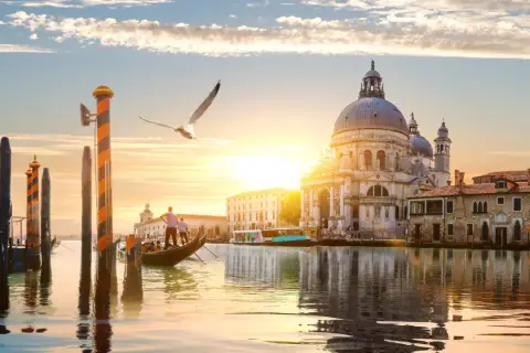 Góndola navega por canal veneciano al atardecer, con la Basílica de Santa Maria della Salute y otras **ciudades de Europa** reflejadas en el agua.