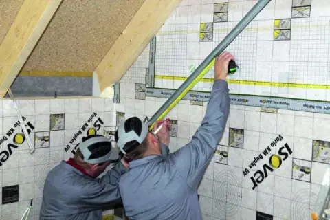 Dwóch mężczyzn pracuje przy montażu grzybków na poddaszu, mierząc i zaznaczając punkty na folii paroizolacyjnej.