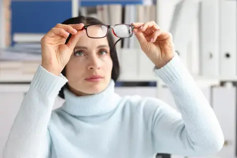 Kobieta w jasnoniebieskim golfie trzyma okulary, myśląc o suplementacji luteiną na oczy dla lepszego widzenia.