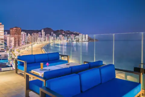 Sofás azules en terraza con vistas a la playa y la ciudad iluminada de Barceló Benidorm Beach al anochecer.