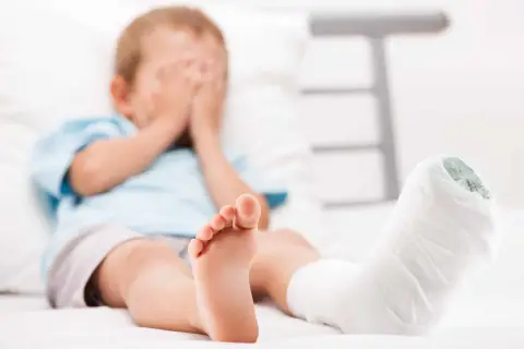 Chłopiec z zabandażowaną nogą, ukrywa twarz w dłoniach. Może to być skutek wypadku lub objaw krzywicy.