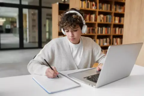 Młoda osoba w słuchawkach uczy się w bibliotece, notując na kartce. Czy nauczanie domowe jest płatne?