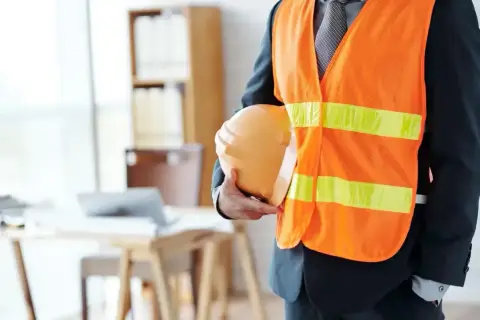 Mężczyzna w kamizelce odblaskowej i garniturze trzyma kask. Pamiętaj o regularnym szkoleniu BHP co ile lat, aby zapewnić bezpieczeństwo.