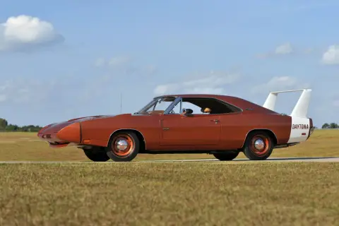 Pomarańczowy Dodge Daytona z charakterystycznym spojlerem i białym pasem z napisem "DAYTONA" na błotniku, zaparkowany na tle nieba z chmurami.