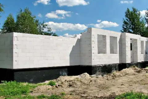 Nowe, białe ściany domu z bloczków betonowych, z otworami na okna i drzwi, stoją na czarnym fundamencie.