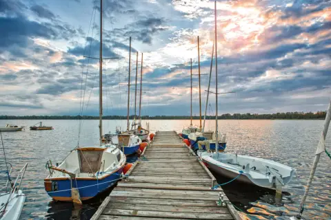 Słoneczny zachód nad pogorią jezioro. Drewniane molo z żaglówkami, chmury odbijające się w wodzie.