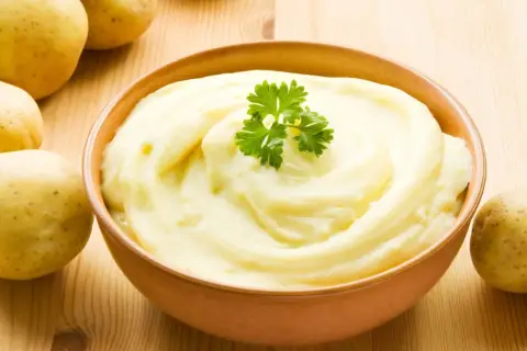 Un tazón de cremoso puré de patatas, decorado con perejil, junto a patatas crudas. ¡Así es como hacer puré de patatas perfecto!