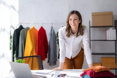 Une chef de produit textile souriante, entourée de vêtements colorés sur un portant, travaille dans son bureau lumineux.