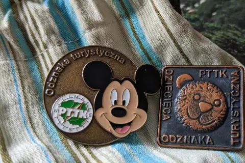 Dwie odznaki turystyczne PTTK: okrągła z Myszką Miki i kwadratowa z misiem.