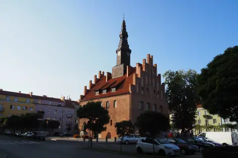 Ratusz w mieście nad jeziorem Skiertąg, z charakterystyczną wieżą zegarową, otoczony kolorowymi kamienicami i zielenią.