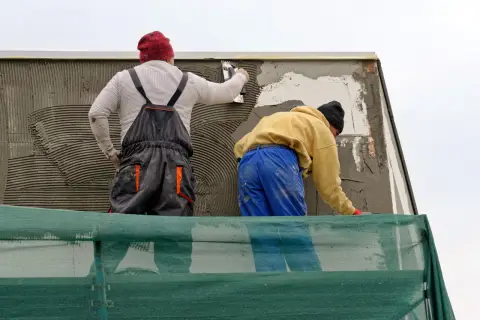 Lavoratori applicano intonaco su un muro esterno, integrando una rete per rinforzo. La rasatura del muro esterno con rete è in corso.