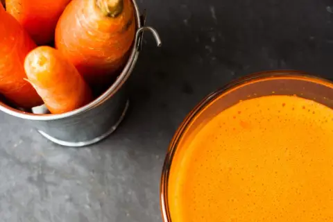 Frische Karotten in einem Eimer neben einem Glas leuchtend orangem Karottensaft. Die Wirkung von Karottensaft ist bekannt für seine Vitamine.
