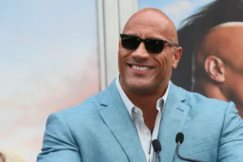 Dwayne "The Rock" Johnson, być może najbogatszy aktor na świecie, w błękitnej marynarce i okularach przeciwsłonecznych, uśmiecha się do kamery.