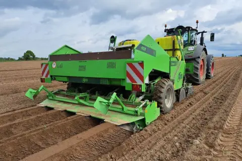 Zielona sadzarka ziemniaków z traktorem na polu. Maszyna wyznacza rzędy, pokazując, na jaką głębokość sadzić ziemniaki.