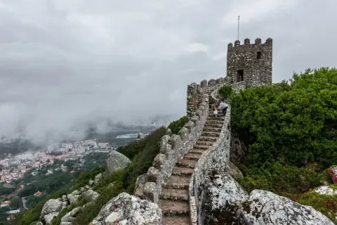 Escaleras de piedra ascienden hacia una torre en el **castillo de Sintra**, con vistas a la ciudad envuelta en niebla.