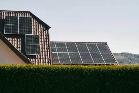 Panele słoneczne na dachu domu, zapewniające czystą energię. W tle las, a na pierwszym planie żywopłot. System jest też zabezpieczony odgromnikiem.