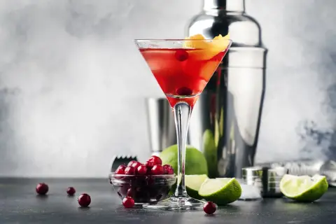 Elegancki Cosmopolitan przepis: czerwony drink w kieliszku, obok żurawina, limonka i shaker. Idealny na wieczór.