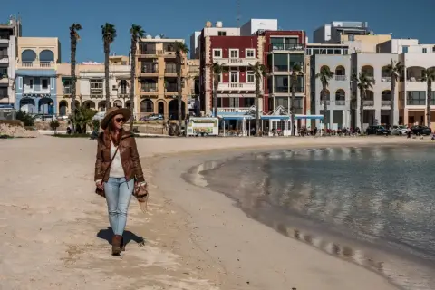 Kobieta spaceruje po piaszczystej plaży w pięknym, słonecznym dniu. W tle kolorowe budynki i palmy tworzą urokliwy krajobraz.