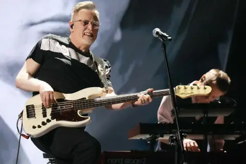 Muzyk gra na białej gitarze basowej, tworząc swoje opus magnum. W tle klawiszowiec pochyla się nad instrumentem.