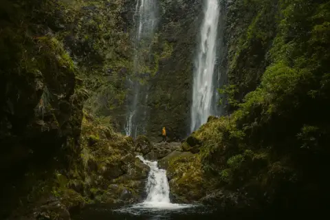 A lone hiker in an orange jacket stands at the base of the majestic Caldeirão Verde waterfall, a breathtaking sight on this lush hike.