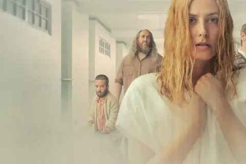 Kobieta z blond włosami i grupa ludzi w korytarzu. Wszyscy wyglądają na zamyślonych, jakby podążali za krzywymi liniami boga.