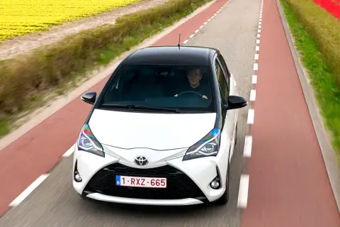 Biały Toyota Yaris Sedan z czarnym dachem jedzie drogą wśród pól żółtych kwiatów i czerwonych tulipanów.