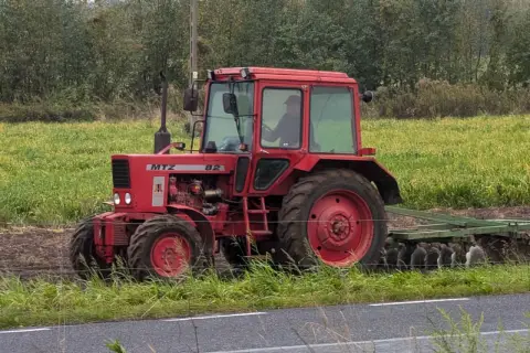 Czerwony ciągnik MTZ Belarus 82 z kabiną pracuje na polu, za nim widać pług.