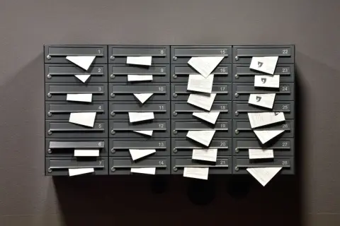 Rząd szarych skrzynek pocztowych z numerami, w których widać wystające białe koperty i kartki.