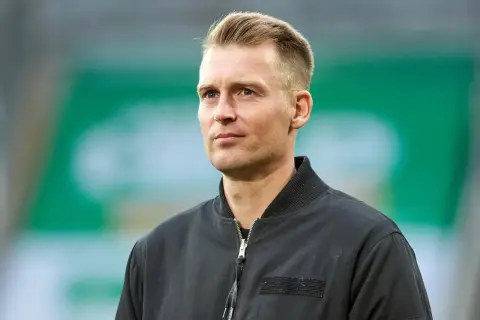 Ein Mann mit blondem Haar und blauen Augen, trägt eine schwarze Bomberjacke. Er blickt nachdenklich zur Seite.