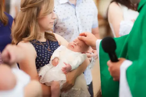 Chrzest dziecka, młoda kobieta trzyma niemowlę, a ksiądz namaszcza je. Czy rodzeństwo może być chrzestnymi?
