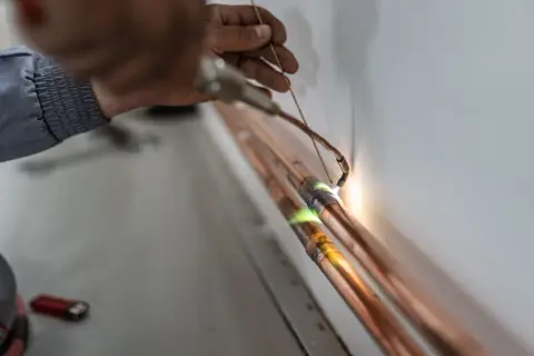 Precyzyjne lutowanie miedzianych rur. Ręka fachowca z palnikiem tworzy trwałe połączenie, zapewniając szczelność instalacji.