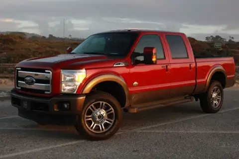 Czerwony Ford F350 King Ranch z brązowym pasem, zaparkowany na tle krajobrazu z wydmami i drewnianym płotem.