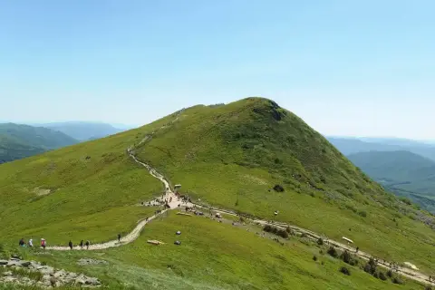 Zielone wzgórza i ścieżka turystyczna prowadząca na szczyt. W oddali widać kolejne pasma górskie, tworząc malowniczy krajobraz, który przypomina o najwyższych szczytach w Polsce.