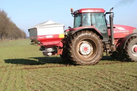Czerwony traktor McCormick z rozsiewaczem na polu. Rozsiewacz rozprowadza saletrę wapniową, kluczową dla zdrowia roślin.