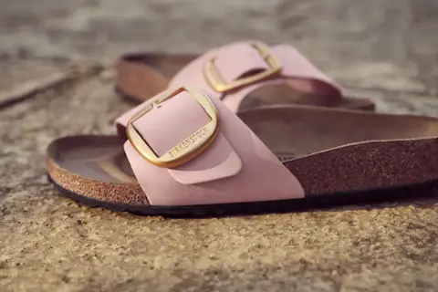 Różowe klapki Birkenstock z złotą klamrą, leżące na kamiennym podłożu.