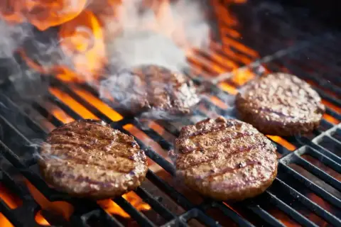 Soczyste burgery z grilla skwierczą na rozżarzonych węglach, otoczone dymem i płomieniami. Idealne na letnie popołudnie.