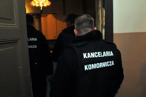Trzech mężczyzn w czarnych kamizelkach z napisem "KANCELARIA KOMORNICZA" wchodzi do pomieszczenia. Prawdopodobnie to postępowanie egzekucyjne.